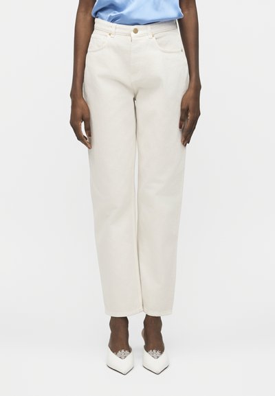 Personne portant un jean blanc taille haute à jambes droites, une chemise bleu clair rentrée, et des chaussures blanches pointues avec un bord en dentelle.