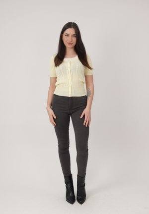 Vrouw met lang donker haar, gekleed in een lichtgeel kort mouwig topje met knopen, donkere strakke spijkerbroek, zwarte enkellaarzen, staand tegen een effen achtergrond.