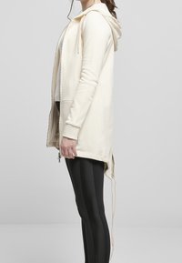 Krämfärgad zip-up hoodie med huva, sidofickor och dragsko. Bärs med svarta leggings, som visar en avslappnad passform och mjukt material.