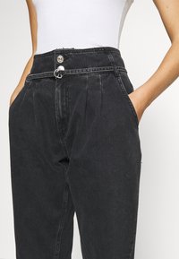 Zwarte denim broek met hoge taille, geplooide voorkant, twee zijzakken en een dubbele knoopsluiting. Voorzien van een riemlus en een lichte vervaging.