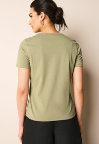 Camiseta de manga corta color verde oliva con una textura suave, cuello redondo y un dobladillo ligeramente curvado, combinada con pantalones negros.