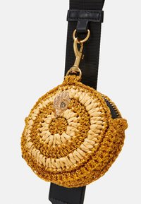 Kulatá tkaná taška ve zlatých a béžových vláknech, s dekorativním kovovým akcentem v podobě pávu, připevněná na černém popruhu s klipovými kovovými prvky.