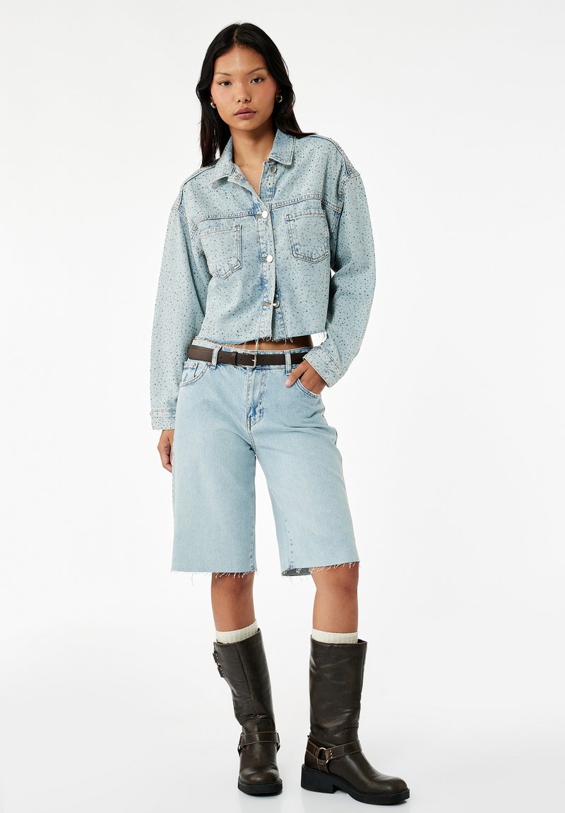 Kvinde iført en lyseblå cropped denimjakke, matchende lange denimshorts, sort bælte, hvide sokker og sorte støvler til midt på læggen, står med hånd i lommen.