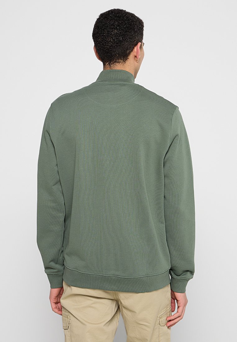 Mężczyzna z krótkimi kręconymi włosami ma na sobie oliwkowy sweter i beżowe spodnie cargo, stoi plecami na jednolitym jasnym tle.