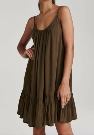 Femme portant une robe marron ample sans manches avec fines bretelles et ourlet volanté, debout, une main sur la hanche et l'autre relâchée.