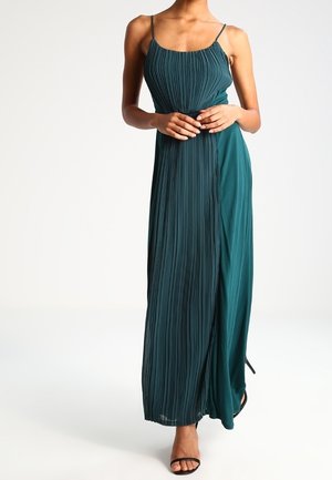 Nainen, joka käyttää hiukan takertumatonta tummaa merensinistä plissoitua maksi-mekkoa, jossa on ohuet hihnat, ja mustia avokkaita kantapäällä, seisoo yksinkertaista taustaa vasten.