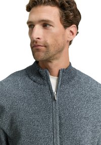 Graue Strickjacke mit Reißverschluss und hohem Kragen, gefertigt aus einem strukturierten Stoff und mit einem silbernen Reißverschluss. Das Design ist lässig und körpernah.