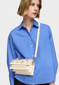 Femme portant une chemise bleue boutonnée et un sac à bandoulière beige orné de décorations florales translucides.