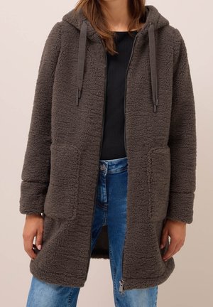 Femme portant une longue veste polaire marron foncé zippée avec capuche, poches avant, haut noir et jean bleu.