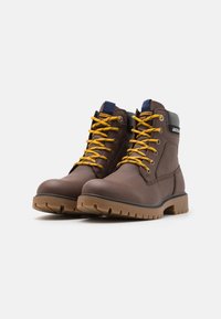 Botas de cuero marrón hasta el tobillo con cordones amarillos, suela de goma y acentos negros. Presentan una etiqueta de logo en la lengüeta y un diseño cosido.