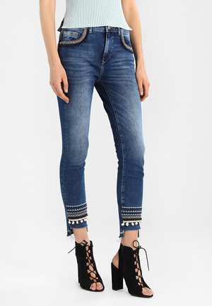 Femme portant un jean en denim bleu avec broderies et bordure à pompons aux chevilles, assorti à des bottes à lacets noires à talons hauts et orteils ouverts.