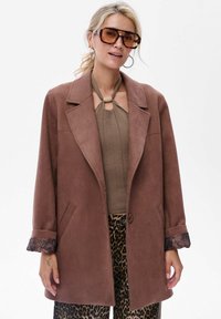 Bruine, getextureerde blazer met notch-revers en een enkele knoopsluiting. De mouwen hebben een gemustreerd stoffen detail. Gecombineerd met een gebreid topje en gemusterde broek.