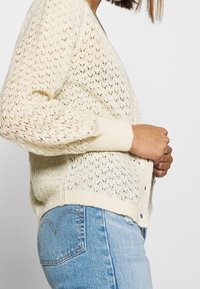 Kremowy kardigan z ażurowym splotem, z dekoltem w serek, długimi rękawami i ściągaczem u dołu. Połączony z jasnobłękitnymi dżinsami.