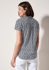 Blusa a maniche corte con motivo di foglie in bianco e nero, dotata di una pinna posteriore bottonata e dettagli arricciati, realizzata in tessuto leggero.