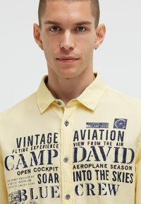 Gelbes Hemd mit Knopfleiste, das mit dunkelblauem Text und luftfahrtbezogenen Grafiken versehen ist. Der Stoff hat eine strukturierte Oberfläche und ein klassisches Kragen-Design.