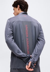 Άνδρας με γκρι μακρυμάνικη μπλούζα Under Armour, χέρια σταυρωμένα πίσω, κόκκινο λογότυπο και κατακόρυφο κείμενο στην πλάτη.