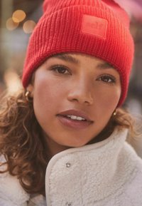 Rød ribbet strikbeanie med en tekstureret finish og et mærketap. Modellen bærer en hvid fleecejakke med sølvfarvede knapdetaljer.