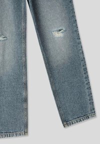 Jeans in denim azzurro chiaro con una vestibilità rilassata, evidenti toppe strappate e orli sfrangiati. Il tessuto ha una consistenza liscia e un sbiadimento sottile.