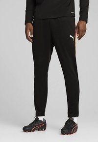 Ανδρας που φορά μαύρο παντελόνι Puma με πορτοκαλί πλαϊνές ρίγες, μαύρα και πορτοκαλί παπούτσια, και λευκές κάλτσες, στέκεται μπροστά σε γκρίζο φόντο.