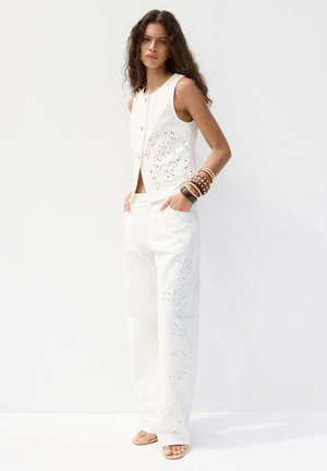 Woman with long curly hair wearing white sleeveless buttoned top and wide-leg pants with cutout patterns, standing with hands in pockets.