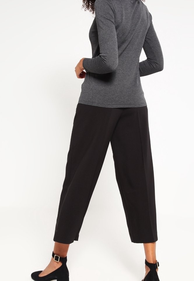 Femme portant un haut gris ajusté à manches longues et un pantalon court noir ample avec des ballerines noires à bride à la cheville, debout, dos tourné.