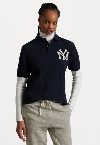 Marineblå polo med hvit New York Yankees-logo, korte ermer, og klassisk krage, lagd over en hvit løpetopp. Grå joggebukser nedenfor.