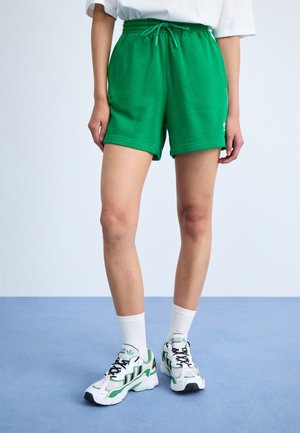 Personne portant un short vert à cordon, des chaussettes blanches et des baskets Adidas blanches et vertes, debout sur un sol bleu devant un fond blanc.