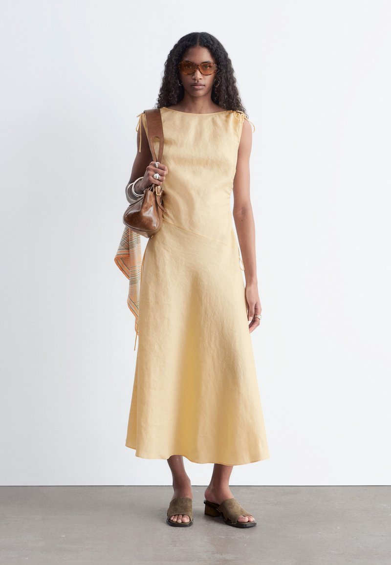 Femme portant une robe beige sans manches, talons ouverts marron, lunettes de soleil ambre, portant un sac à main en cuir marron avec une écharpe à motifs.