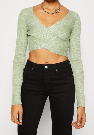Female torso wearing a light green long-sleeve cropped wrap sweater, black high-waisted jeans, and a silver heart-shaped chain necklace.