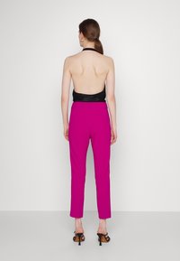 Vrouw met bruin haar in een paardenstaart, gekleed in een zwarte haltertop met een open rug, felroze hoge taillebroek en zwarte hoge hakken, staande met haar rug naar de kijker.