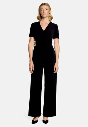 Vrouw met blond haar die een zwarte jumpsuit met V-hals, korte mouwen en wijde pijpen draagt, staand met ontspannen armen langs het lichaam.
