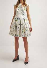 Kleita ar ziedu rakstu izgatavota no krēmkrāsas auduma, ar īsām piedurknēm, izteiktu jostasvietu un sakrautu svārku. Saskaņota ar melniem augstiem papēžiem.