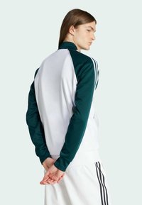Zelená a bílá sportovní bunda se zipem, kontrastní design rukávů, žebrovaný límec a tři proužky podél každého rukávu. Hladká textura.