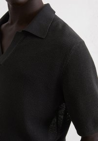 Polo noir texturé à manches courtes avec un col en V, présentant des détails en tissage et une coupe légèrement ample.