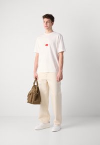 Lyse beige t-shirt med et rødt grafisk motiv i midten, parret med cremede bukser og hvide sneakers. Brun håndtaske med fremtrædende hardware detaljer.