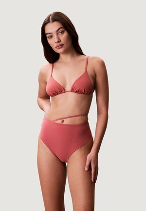 Młoda kobieta z długimi brązowymi włosami, ubrana w dwuczęściowy bikini w kolorze przygaszonego różu z wysokim stanem, stoi na tle jednolitego jasnego tła.