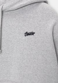 Harmaa hupullinen collegepaita, jossa on pehmeä tekstuuri ja vasemmassa rinnassa musta kirjailtu logo. Hupussa on nyörit.