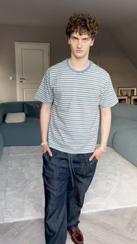 Šviesiai mėlynas ir pilkas dryžuotas trumpomis rankovėmis marškinėlis, derinamas su tamsiomis laisvos formos kelnėmis. Modelis stovi atsipalaidavęs, rankas įsidėjęs į kišenes.