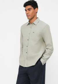 Camicia button-up grigio chiaro in un tessuto testurizzato con un logo verde sulla tasca del petto. Presenta un colletto classico e una forma aderente.