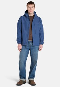 Blå vattentät jacka med huva, dragkedja fram, elastiska ärmslut och två framfickor. Buren över en brun topp, kombinerad med blå jeans och bruna skor.