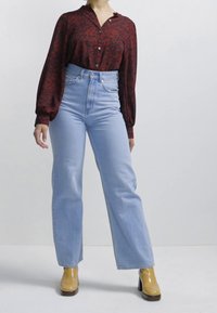 Camisa de botão vermelha com flores e mangas balão, combinada com calças de ganga claras de cintura alta e pernas largas, e botas amarelas de salto.