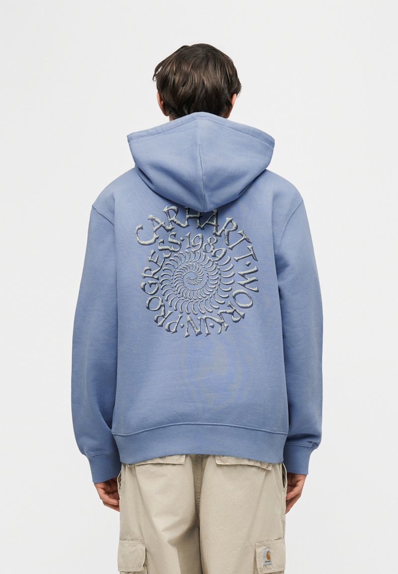 Persona vistiendo una sudadera con capucha azul con texto en espiral "Carhartt Work In Progress 1989" en la espalda, combinada con pantalones cargo beige.