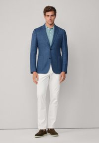 Modrý saka na míru s klopovými límci, doplněný světle modrou polokošilí. Bílé kalhoty a hnědé slip-on boty dotvářejí celkový vzhled.