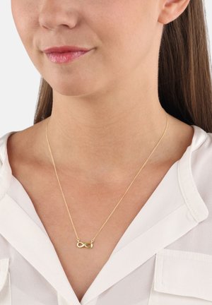 Femme portant un délicat collier en or en forme d'infini avec un chemisier blanc à col en V, montré de la mâchoire à la partie supérieure de la poitrine.