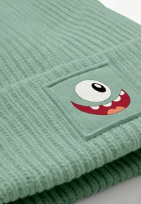 Mint green knitted beanie featuring a square patch with a cartoonish face, notable for its large eye and wide smiling mouth.