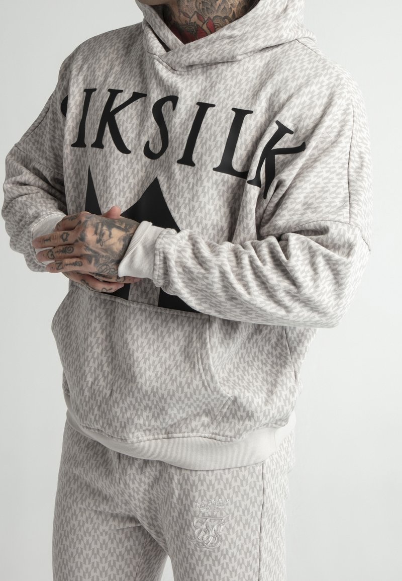 Personne tatouée dans un survêtement gris clair à motifs portant l’inscription “SIKSILK”, mains jointes, seuls le haut du corps et les jambes sont visibles, fond neutre.