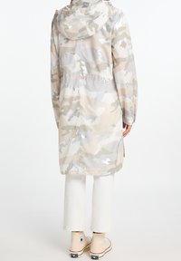 Płaszcz przeciwdeszczowy w beżowych i szarych odcieniach z kapturem, sznurkiem ściągającym w talii oraz lekkim, błyszczącym materiałem. Zestawiony z białymi spodniami i butami.