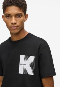 Jaunais vīrietis valkā melnu T-kreklu ar baltu izkropļotu "K" logotipu un "Karl Lagerfeld Jeans" uzrakstu uz krūšu pret vienkāršu gaišu fonu.