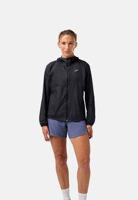 Veste noire légère avec capuche, fermeture éclair et poignets élastiques. Portée sur un short bleu, avec un logo discret sur la poitrine.