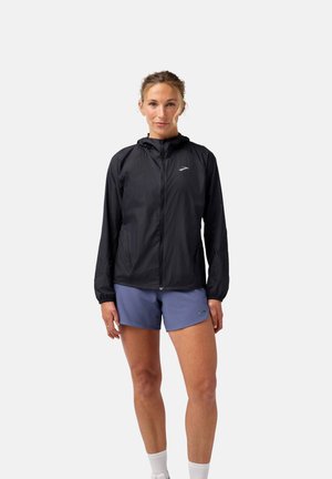 Leichte schwarze Jacke mit Kapuze, Reißverschluss und elastischen Bündchen. Über blauen Shorts getragen, mit einem dezenten Logo auf der Brust.